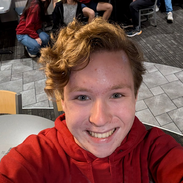 Left: Luke Baker poses in safety goggles, his red hair peeking out from the visor of the white helmet that covers his head, mouth, and chin.  Right: Luke Baker takes a selfie with a group of smiling students in a restaurant.