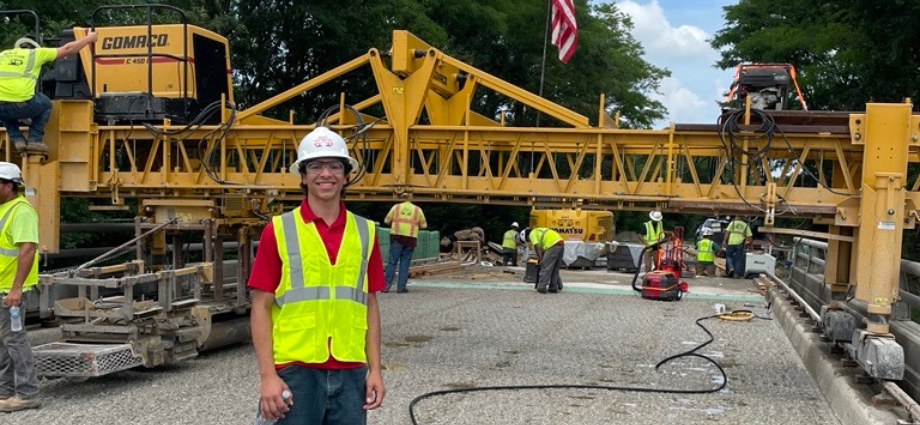 Egan Walsh in a hard hat and safety vest by large construction machinery.