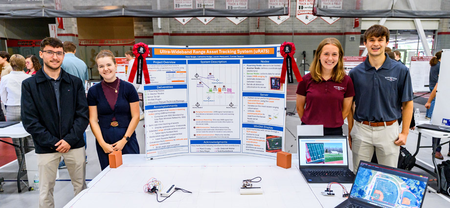 Students pose with award ribbons behind a table, which displays their project on a trifold board and a laptop.