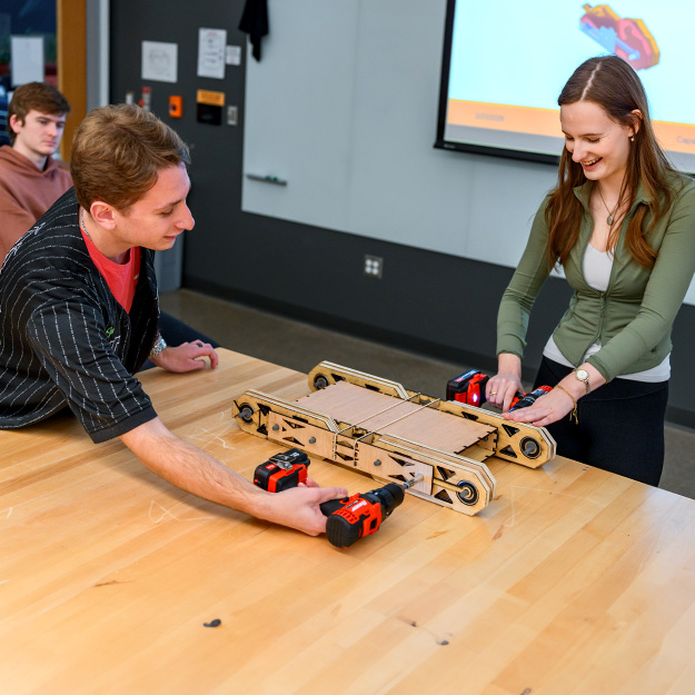 A faculty and students work together in a classroom lab, assembling and testing a cardboard prototype device on a table.