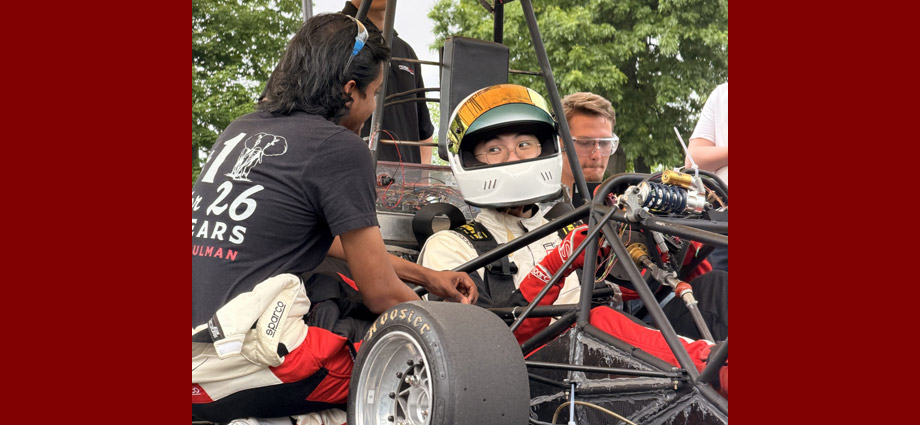 A studnet sits in a race vehicle wearing a helmet and talking to another student.