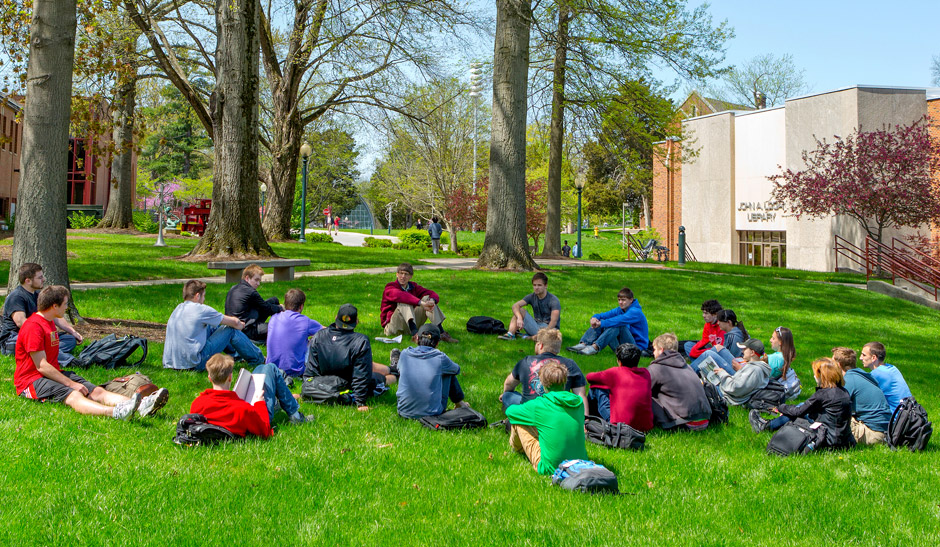 Students seated in the grass in the Root Quadrangle listening to a professor, who is also seated.