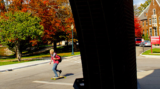 A student riding a skateboard on the road with Deming Hall in the background. 