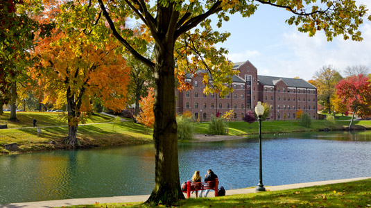 Two female students sit on a bench next to Speed Lake on a sunny fall day. Percopo Hall dormitory is in the background across the lake.
