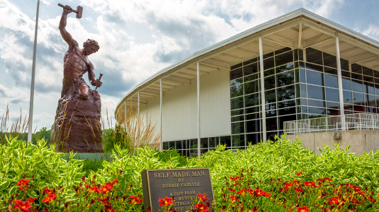 Self-made Man sculpture in the foreground amid green plants and red flowers with the Sports and Recreation Center in the background.