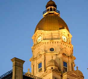 Image shows the Vigo County Courthouse with its dome and large clocks. The dome and tower are illuminated against a dark blue evening sky.
